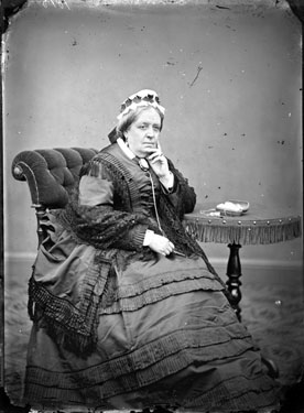 Portrait of a woman in 'Queen Victoria' pose: she has a cameo brooch, lace work in a case, eye glasses on a string and is inscribed '1875'